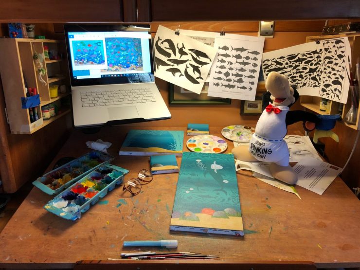 This is a photo of a painting desk, with canvases and paints. On the desk is a stuffed toy Opus, a penguin character from the comic strip, Bloom County